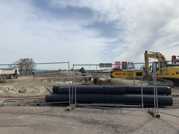 Großer Haufen schwarzer Rohre auf einer Baustelle, umgeben von einem Metallzaun, mit einem Bagger rechts und Fahrzeugen auf der Straße links, Bäume und Strommasten im Hintergrund.