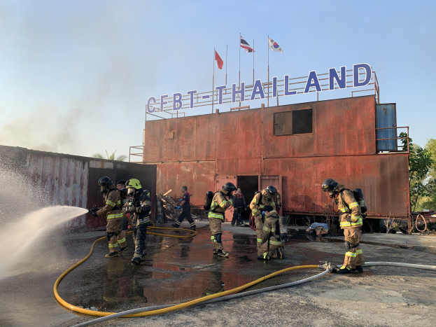 Feuerwehrleute in Helmen sprühen Wasser auf ein Gebäude mit Text, während Flaggen, Bäume und ein klarer Himmel im Hintergrund zu sehen sind.