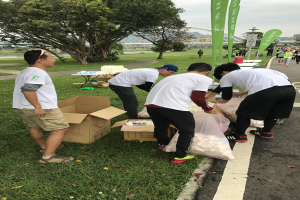 Gruppe von Menschen an einer Straße mit verstreuten Kartons und Flaschen, umgeben von Bäumen und Gebäuden, die an einem Charity-Lauf unter einem klaren blauen Himmel teilnehmen.