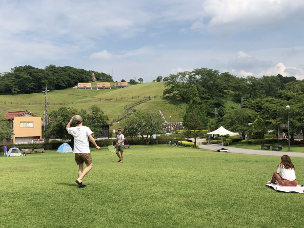 Eine Gruppe von Menschen, die Badminton in einem Park spielt, mit Zelten, Straßenmöbeln und Gebäuden im Hintergrund.