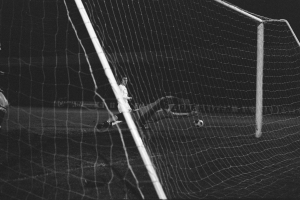 Ein Fussballer in Schwarz-Wei\ss-Fotografie macht eine Parade, mit einem Ball in der Luft und einem Netz im Hintergrund.