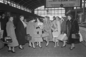 Schwarzes und weißes Bild von Frauen, die an einem Bahnhof stehen, jede hält eine Tasche, mit einem Zug links und Geländern und einer Tafel rechts.
