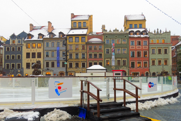 Eisbahn in einem Stadtplatz umgeben von Gebäuden, Treppen, Geländern, Informationsschildern, schneebedecktem Boden, Laternen, Bäumen und einem Himmel.