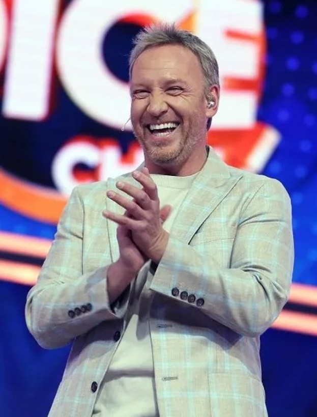 Ein Mann in einem Anzug, der auf einer Bühne klatscht, mit 'The Voice'-Zeichen im Hintergrund.