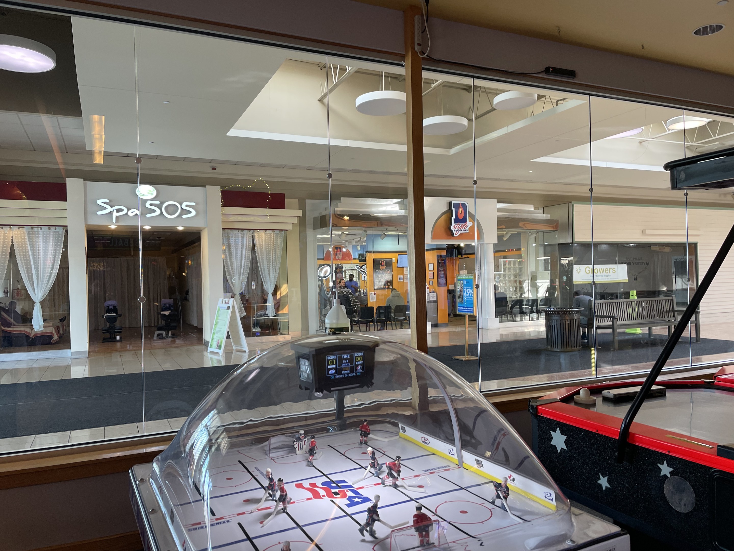 Eine Eishalle in einem Einkaufszentrum, umgeben von Glaswänden mit Menschen, Möbeln und Beschriftungen, die durch sie hindurch sichtbar sind, und einem Air-Hockey-Tisch auf der rechten Seite.