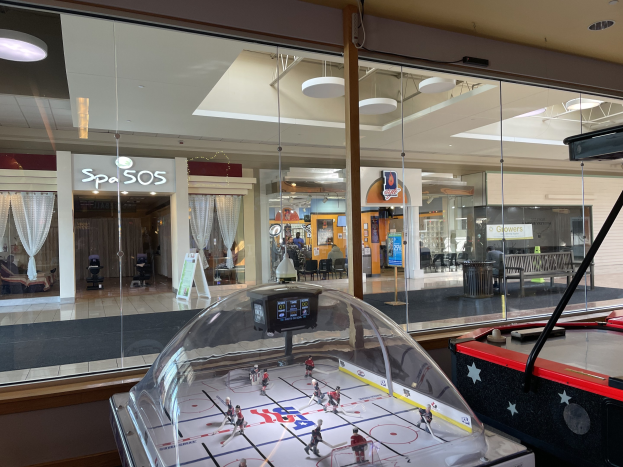 Eine Eishalle in einem Einkaufszentrum, umgeben von Glaswänden mit Menschen, Möbeln und Beschriftungen, die durch sie hindurch sichtbar sind, und einem Air-Hockey-Tisch auf der rechten Seite.