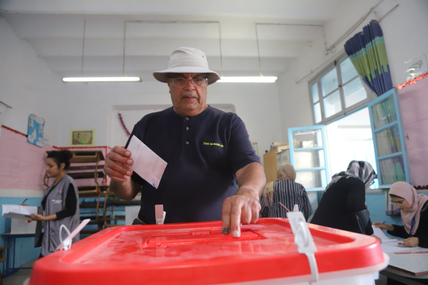 Ein Mann mit Hut stimmt bei einer Wahllokal ab, vor einer roten Wahlurne stehend, mit einem Stück Papier in der Hand, während andere an Tischen sitzen und schreiben, in der Nähe eines Fensters mit einem Vorhang.