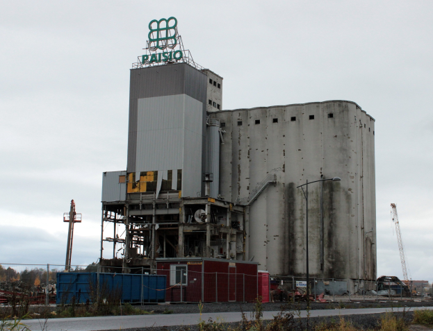 Großes Zementgebäude neben einer Straße, umgeben von Vegetation, einem Zaun, Strommasten und Straßenlaternen, mit Bäumen und einem klaren blauen Himmel im Hintergrund.