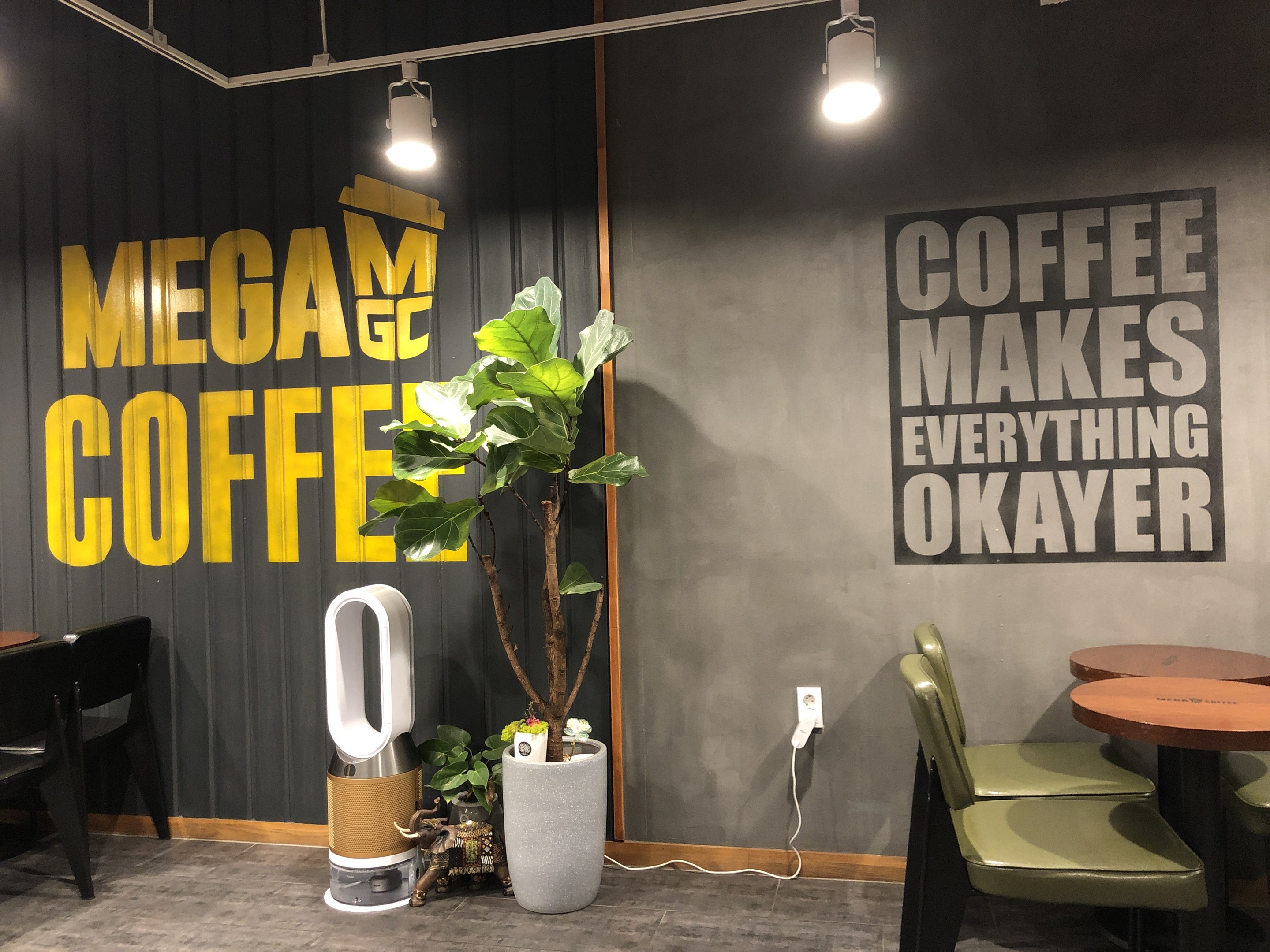 Außenansicht eines Kaffees mit dem Namen "Mega Coffee" mit Tischen, Stühlen, einem Bodenständer, Deckenleuchten und einem Topf mit einer Pflanze in Singapur.