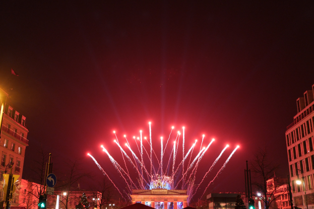 Eine nächtliche Stadtstraße in Berlin mit dem Brandenburger Tor in der Mitte, umgeben von beleuchteten Gebäuden, Bäumen, Pfählen, Verkehrszeichen, Schildern und Fahnen, mit Feuerwerk am Himmel über dem Tor.