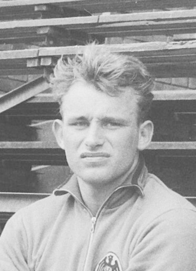 Schwarzes und weißes Foto eines Mannes in einer Jacke, der vor gestapelten Holzplanken steht, identifiziert als legendärer deutscher Fußballspieler, der im Alter von 89 Jahren starb.