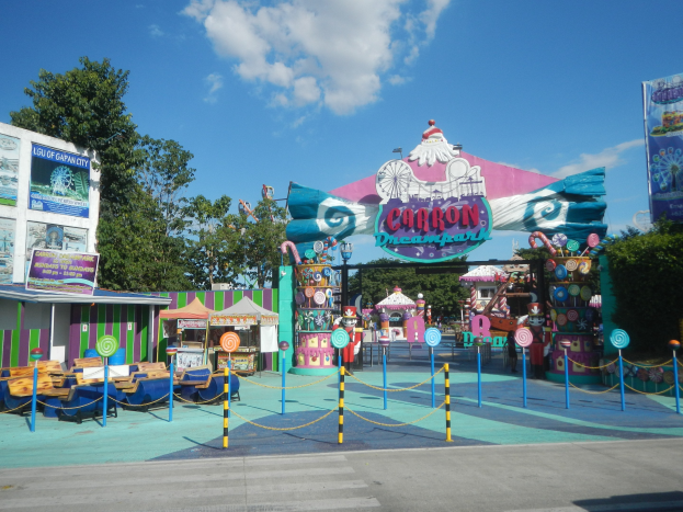 Kirmesfahrgeschäft am Parkeingang mit Straße darunter, eingezäuntem Bereich, Buden, Bannern, Bäumen und bewölktem Himmel.