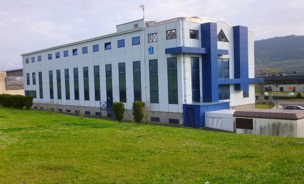 Großes weißes und blaues Gebäude mit Fahrzeugen, Masten und Bergen im Hintergrund, auf einem grünen Feld unter einem klaren blauen Himmel stehend.