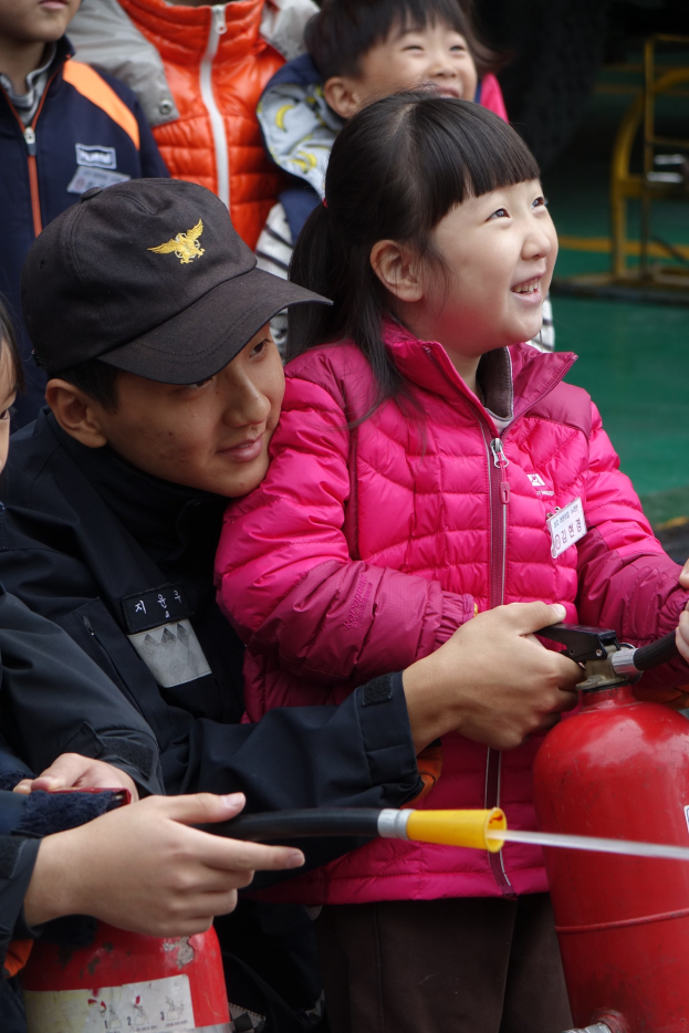 Kinder in einer Schulumgebung um einen roten Feuerhydranten versammelt, einer hält ein Rohr, mit verschiedenen Gegenständen im Hintergrund.