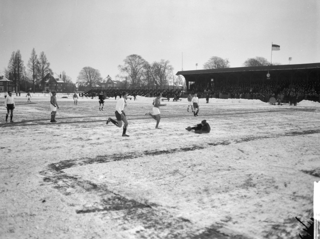 Schwarzes Bild von Menschen, die auf einem schneebedeckten Fußballfeld mit Bäumen, Häusern, Pfählen, Fahnen und einer Hütte im Hintergrund Fußball spielen.