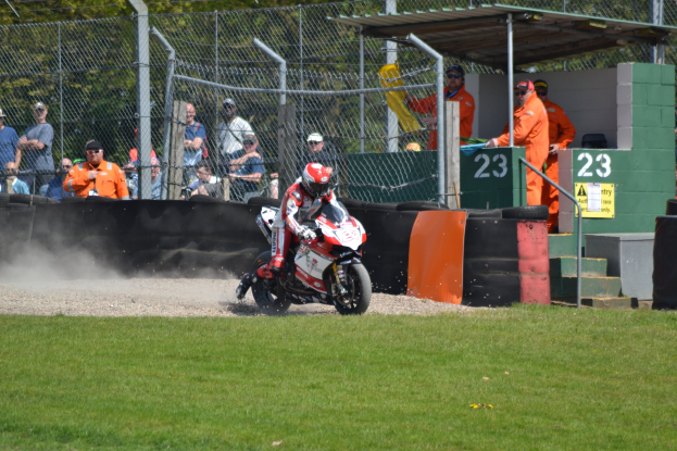 Ein Motorradfahrer in Helm und Handschuhen fährt über ein grasbewachsenes Feld, mit Zuschauern in Kappen und Schutzbrillen, einem eingezäunten Bereich mit Bäumen, Treppen mit Geländern und einem nummerierten Schuppen mit Texttafeln im Hintergrund.