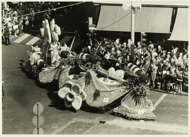 Ein Schwarz-Weiß-Foto eines mit Blumen geschmückten Umzugswagens, mit einer Gruppe von Menschen auf der Straße und einem Pfahl im Hintergrund.