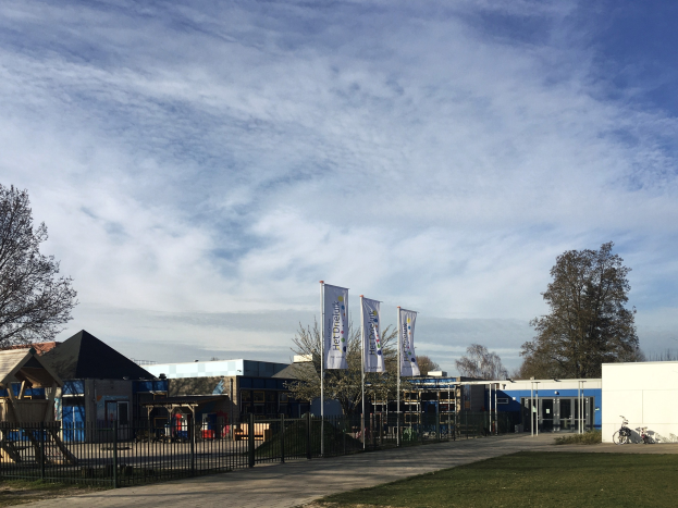 Gro├če Geb├Ąude mit einem gras├Ąhnlichen Bereich davor, umgeben von einem Metallzaun, mit einem Weg, B├Ąumen, Pfosten, Bannern, einem geparkten Fahrrad, n├Ąheren H├Ąusern und einem bew├Âlktem Himmel, als das neue Kinderspielgel├Ąnde an der Schule identifiziert.