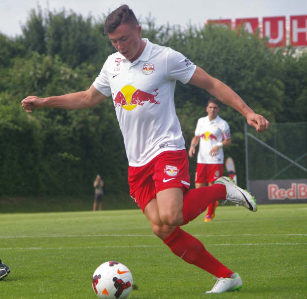 Ein professioneller Fußballspieler in einem roten Trikot tritt einen Ball auf einem grünen Feld mit Bäumen und einem klaren blauen Himmel im Hintergrund, wobei "RB Leipzig" auf einer Tafel zu sehen ist.