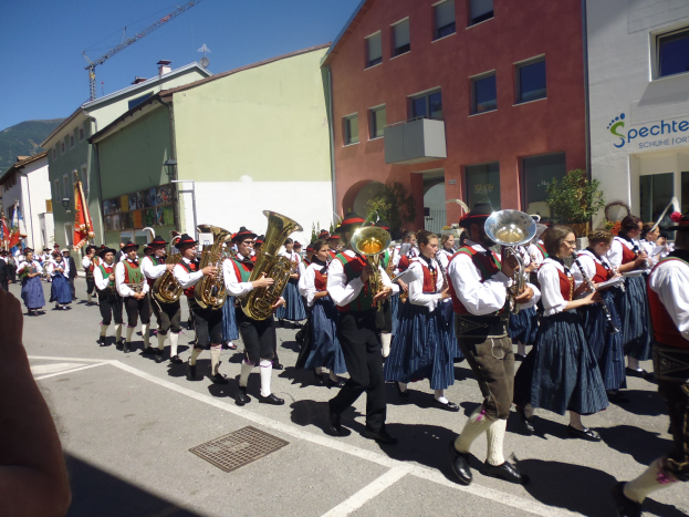 Menschen in traditioneller bayrischer Tracht spielen Musikinstrumente, während sie eine Straße entlanggehen, die von Gebäuden gesäumt ist, einige halten Fahnen, mit einem Hügel und einem blauen Himmel im Hintergrund.