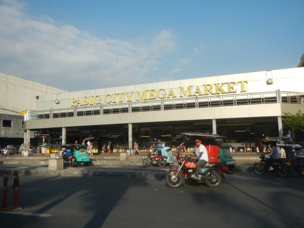 Eine belebte Straßenszene auf dem Pasig City Mega Market mit Fahrzeugen und Fußgängern, mit einem Gebäude mit Text im Hintergrund unter einem bewölkten Himmel.