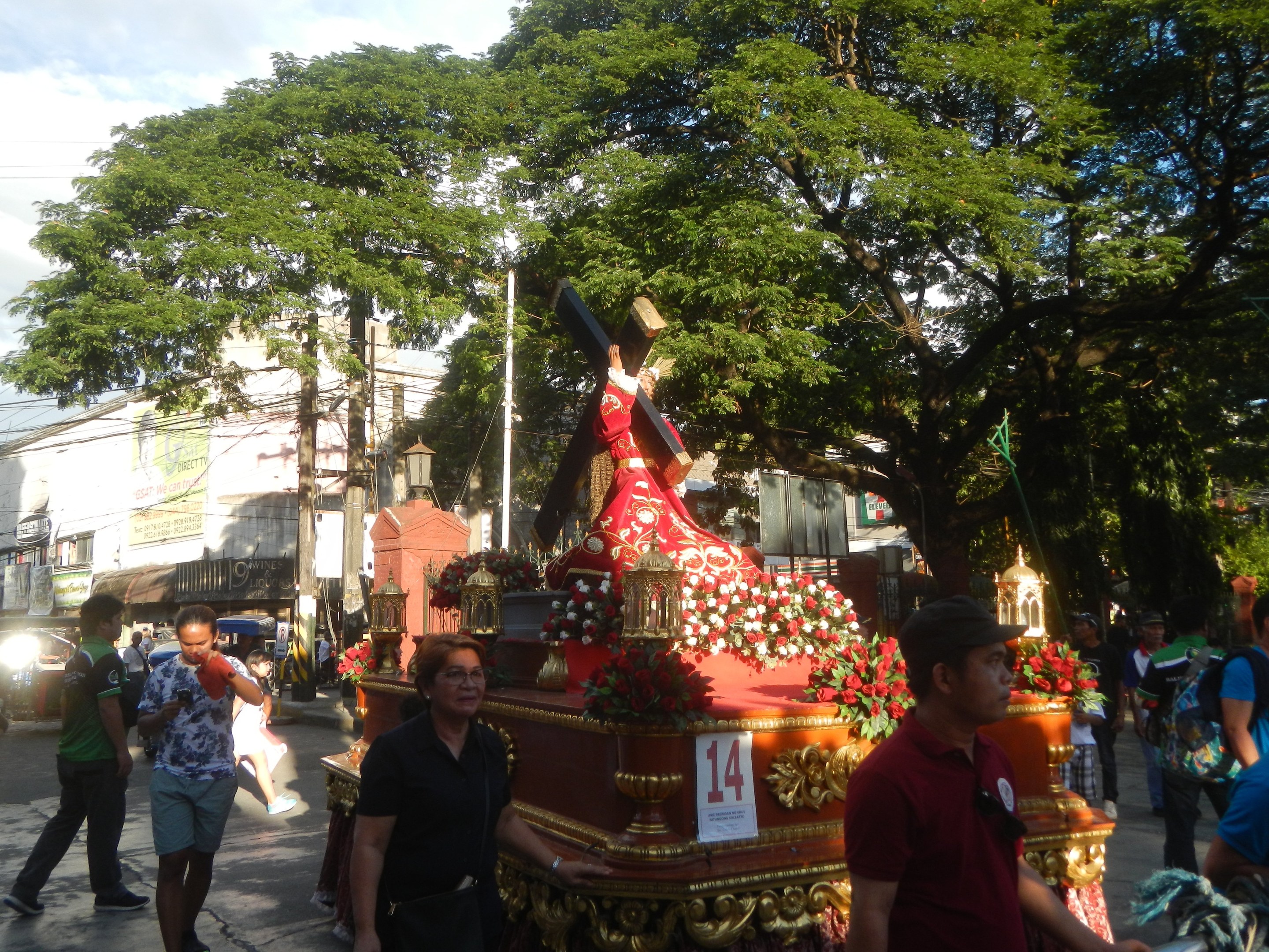 Eine Gruppe von Menschen, die in einer Prozession an einer Jesus-Statue vorbeigehen, mit Bäumen, Gebäuden, Strommasten und Fahrzeugen im Hintergrund, unter einem bewölkten Himmel.
