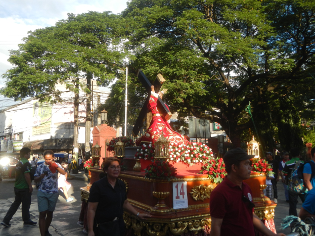 Eine Gruppe von Menschen, die in einer Prozession an einer Jesus-Statue vorbeigehen, mit Bäumen, Gebäuden, Strommasten und Fahrzeugen im Hintergrund, unter einem bewölkten Himmel.