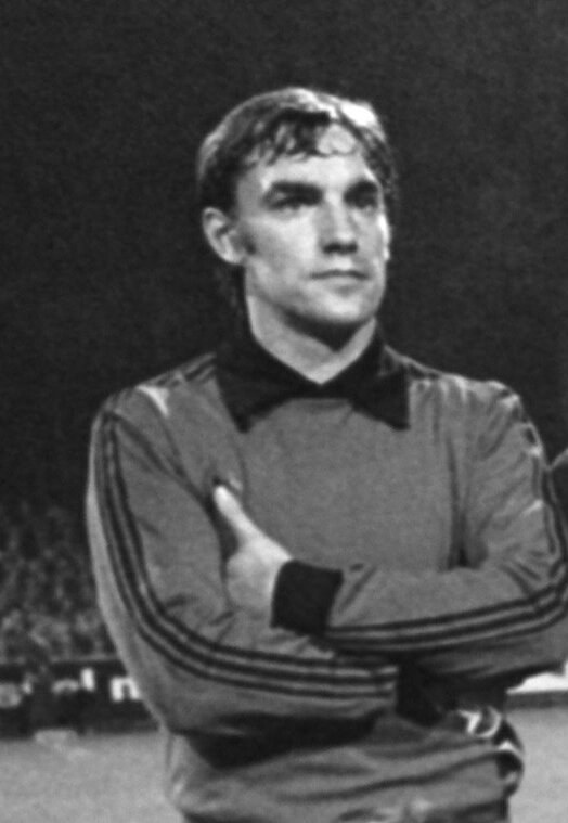 Schwarzes und weißes Foto eines Mannes auf einem Fußballfeld mit verschränkten Armen, umgeben von einer Gruppe von Sitzenden im Hintergrund.