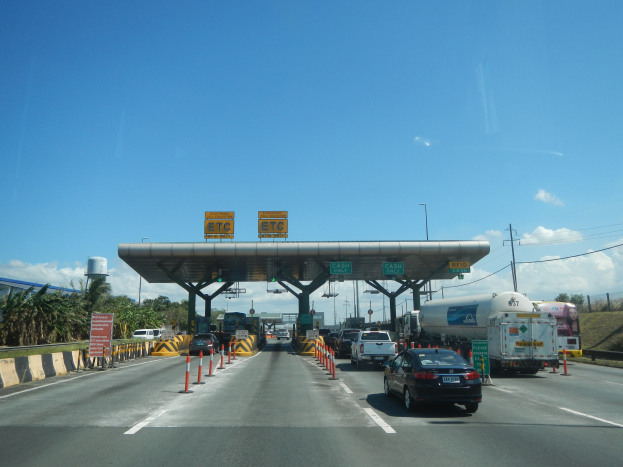 Mautstelle auf einer Autobahn mit Autos, Verkehrskegeln, Informationsschildern, Laternenmasten, Strommasten, Bäumen, einer Hütte und einem bewölkten Himmel.