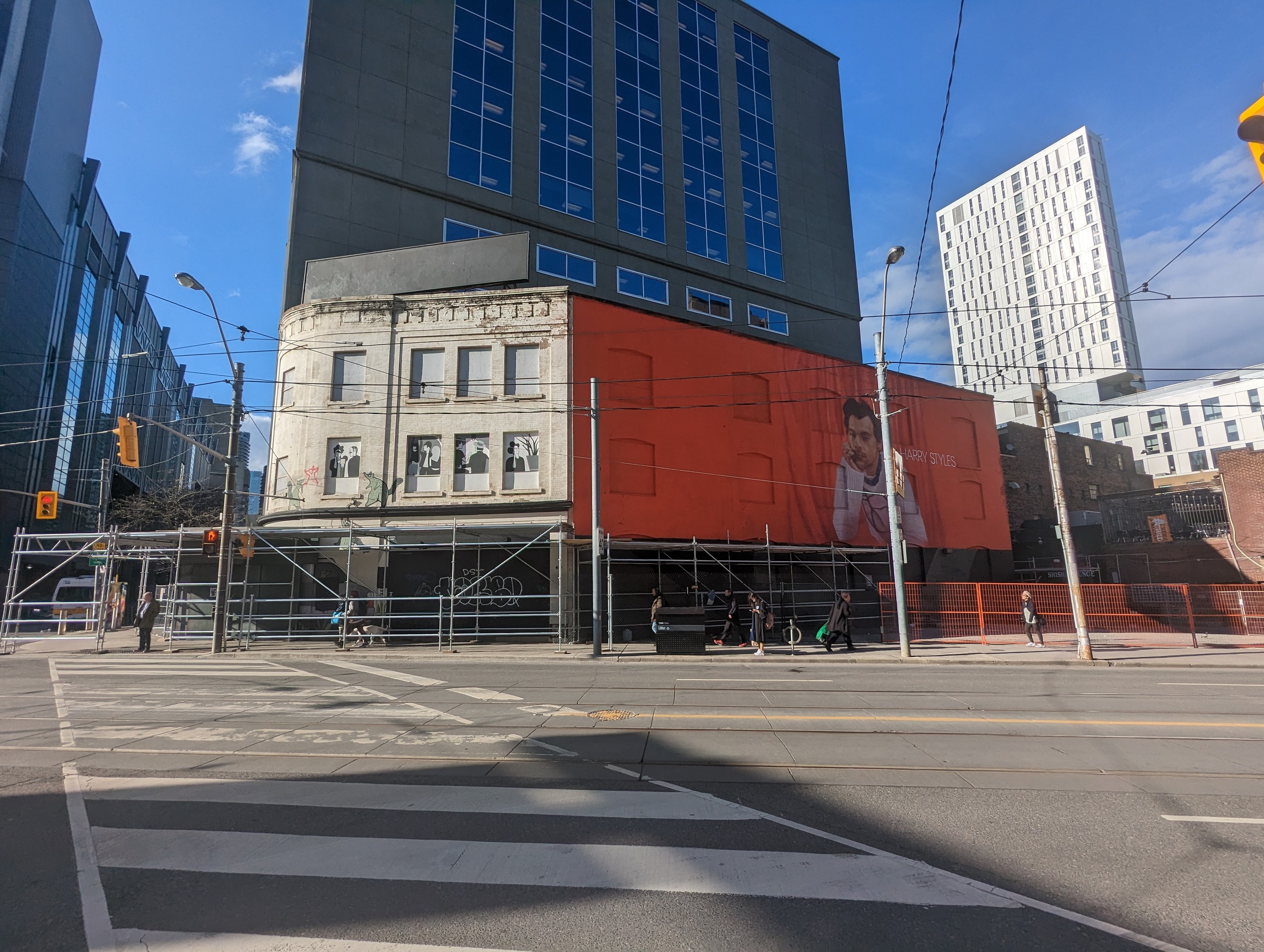 Ein großes Gebäude mit einer Wandmalerei an der Seite, umgeben von Straßen- und Verkehrsinfrastruktur, Fahrzeugen, Fußgängern, Grills, Bäumen und einem bewölkten Himmel, identifiziert als das Museum of Contemporary Art in New York.