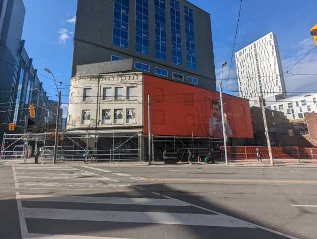 Ein großes Gebäude mit einer Wandmalerei an der Seite, umgeben von Straßen- und Verkehrsinfrastruktur, Fahrzeugen, Fußgängern, Grills, Bäumen und einem bewölkten Himmel, identifiziert als das Museum of Contemporary Art in New York.