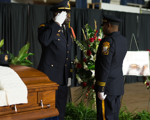 Zwei Polizeibeamte in Uniform stehen neben einem mit weißem Tuch bedeckten Sarg.