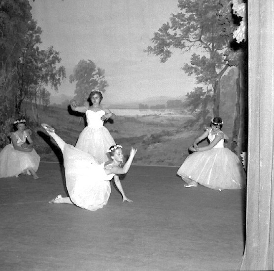 Vier junge Balletttänzerinnen in weißen Kleidern tanzen auf einer Bühne mit einer gemalten Kulisse aus Bäumen und Himmel, in Schwarz-Weiß.
