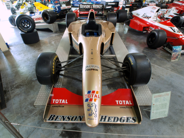 Rennwagen in einem Motorsportmuseum ausgestellt, getrennt von Zuschauern durch einen Metallzaun, mit informativem Text im Hintergrund.