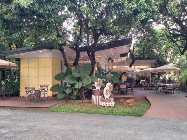 Outdoor-Café mit Tischen, Stühlen und Sonnenschirmen in einer Parkumgebung, umgeben von Bäumen und Pflanzen, mit einer Straße und einem Gebäude im Hintergrund.