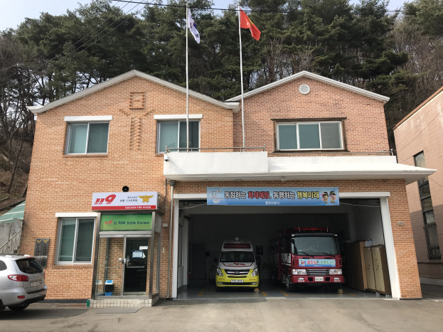 Feuerwache mit zwei geparkten Feuerwehrautos, umgeben von Gebäuden, Flaggen, Informationsschildern, Bäumen und einem klaren blauen Himmel.
