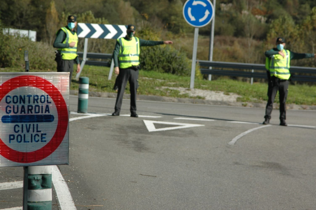 Drei Polizeibeamte in grünen Jacken und schwarzen Hosen stehen an einer Straßenböschung neben Bäumen und einem Geländer, mit einem Verkehrsschild und einem Schuh im Vordergrund.