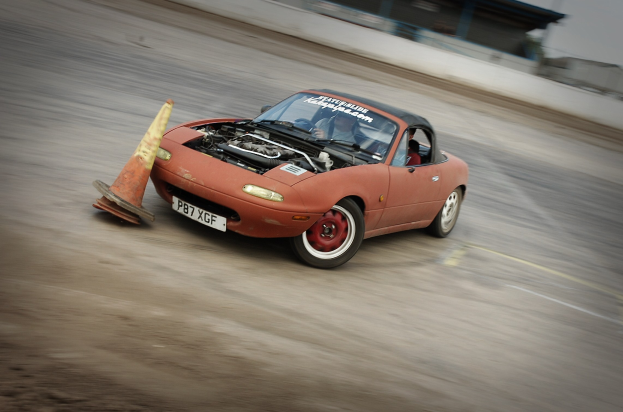 Roter Wagen auf einer Rennstrecke mit einem Verkehrskegel vorne, Hintergrund durch hohe Geschwindigkeit unscharf.