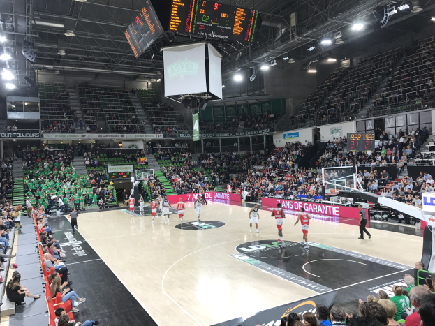 Basketballspiel in einer großen Arena mit Publikum, Anzeigetafeln mit "Asvel vs Galatasaray" und Deckenbeleuchtung und Bildschirm.