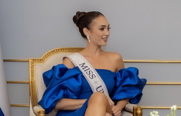 Frau in einem blauen Kleid mit einer 'Miss Universe 2019'-Schärpe, die auf einem Stuhl sitzt und von Blumen umgeben ist.