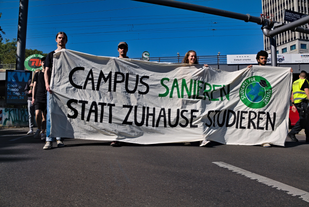 Gruppe von Menschen, die eine Straße entlanggehen und ein Banner mit der Aufschrift "Campus Sanien Statt Zuhause Studieren" halten, mit Gebäuden, Bäumen, Polen, Drähten und einem klaren blauen Himmel im Hintergrund.