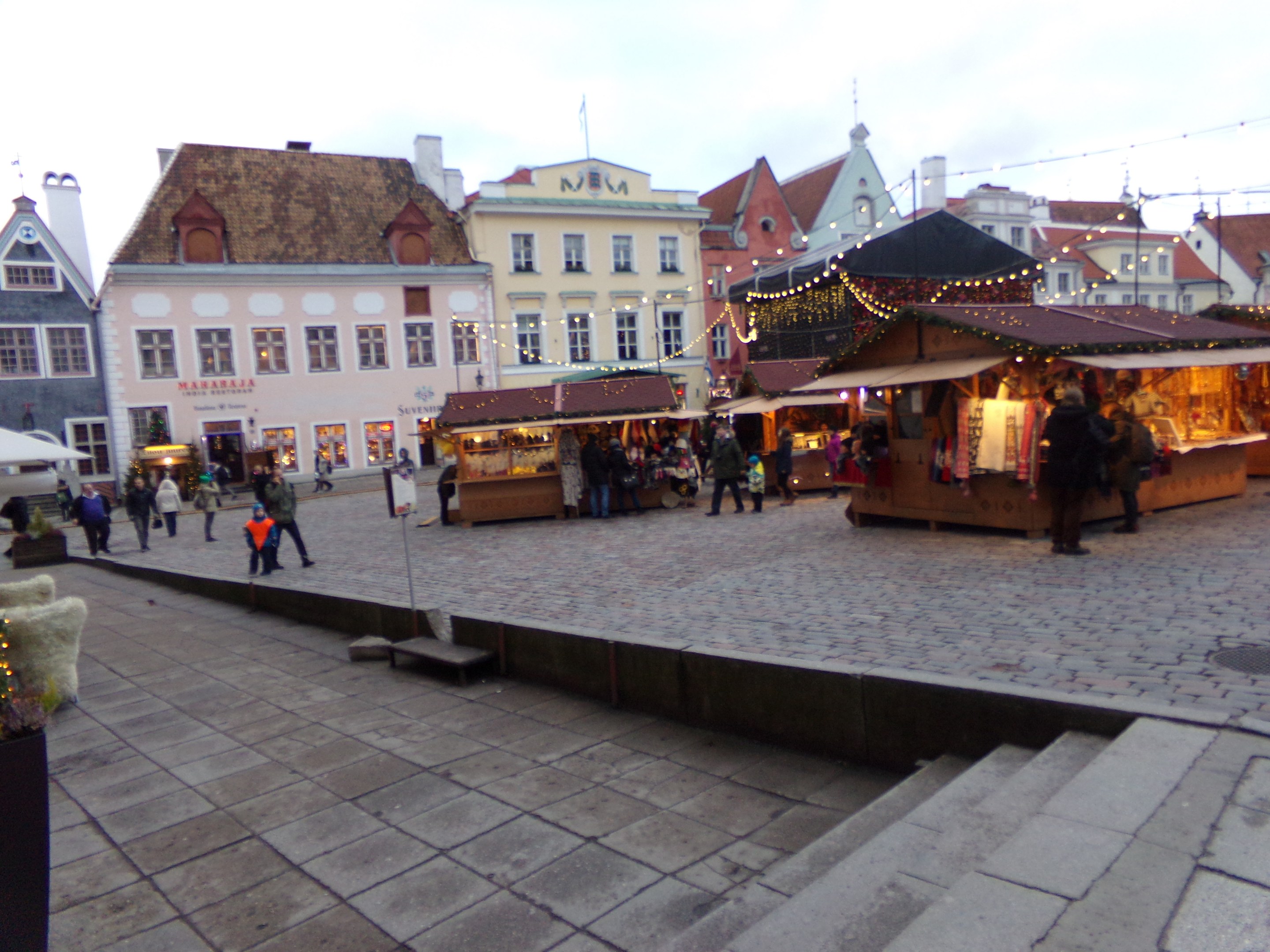 Ein belebter Weihnachtsmarkt in Tallinn, Estland mit Menschen um dekorierte Stände, festlich geschmütkte Gebäude im Hintergrund, bewölktem Himmel, Treppen fürhende zum Markt und Topfpflanzen.