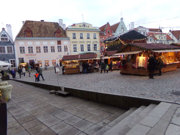 Ein belebter Weihnachtsmarkt in Tallinn, Estland mit Menschen um dekorierte Stände, festlich geschmütkte Gebäude im Hintergrund, bewölktem Himmel, Treppen fürhende zum Markt und Topfpflanzen.