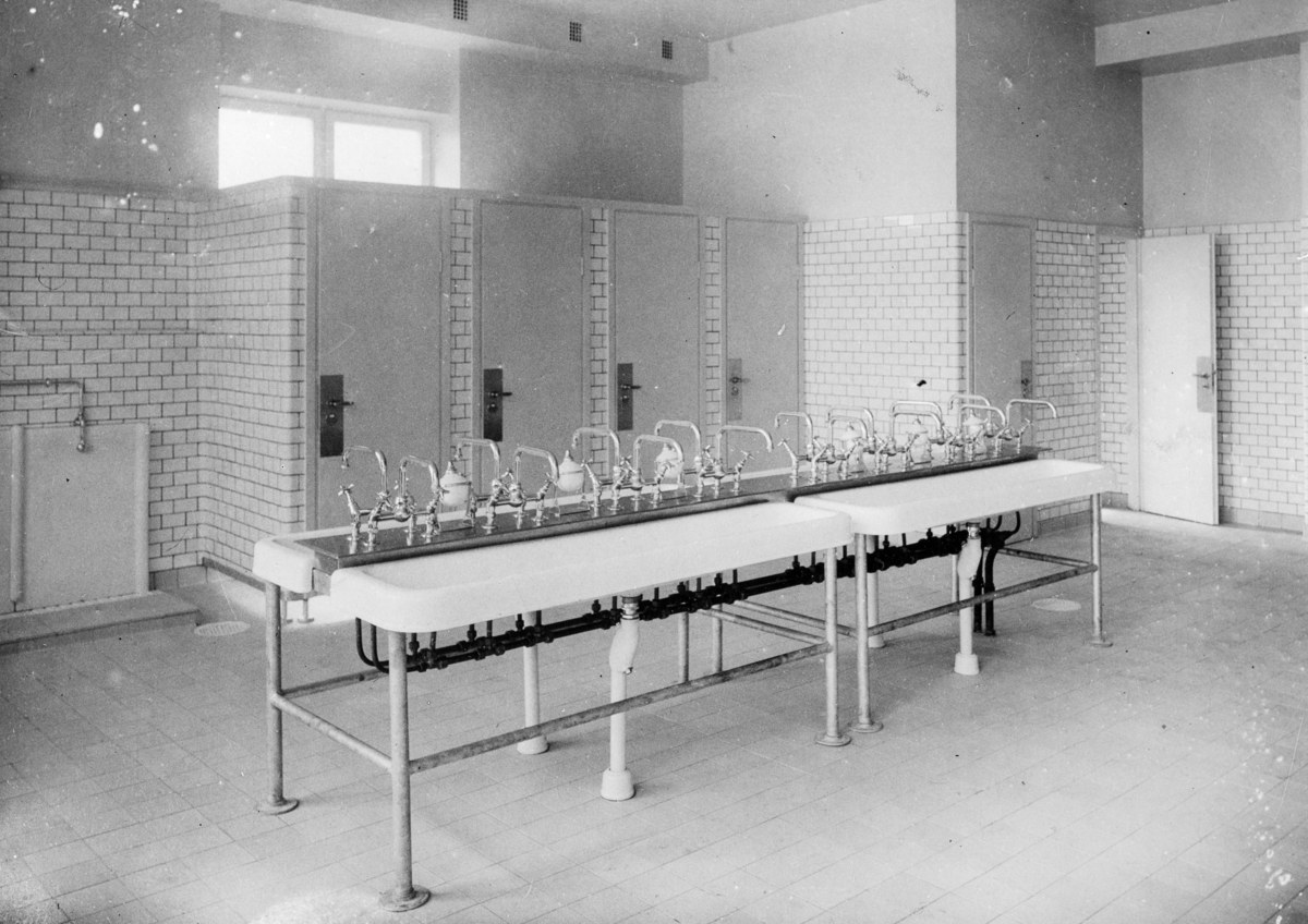 Schwarz-weißes Foto einer öffentlichen Toilette mit einer Reihe von Waschbecken, Wasserhähnen, Wänden, Türen, Fenstern und einem gefliesten Boden, mit verschiedenen Gegenständen, die herumliegen.