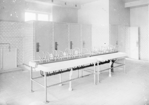 Schwarz-weißes Foto einer öffentlichen Toilette mit einer Reihe von Waschbecken, Wasserhähnen, Wänden, Türen, Fenstern und einem gefliesten Boden, mit verschiedenen Gegenständen, die herumliegen.