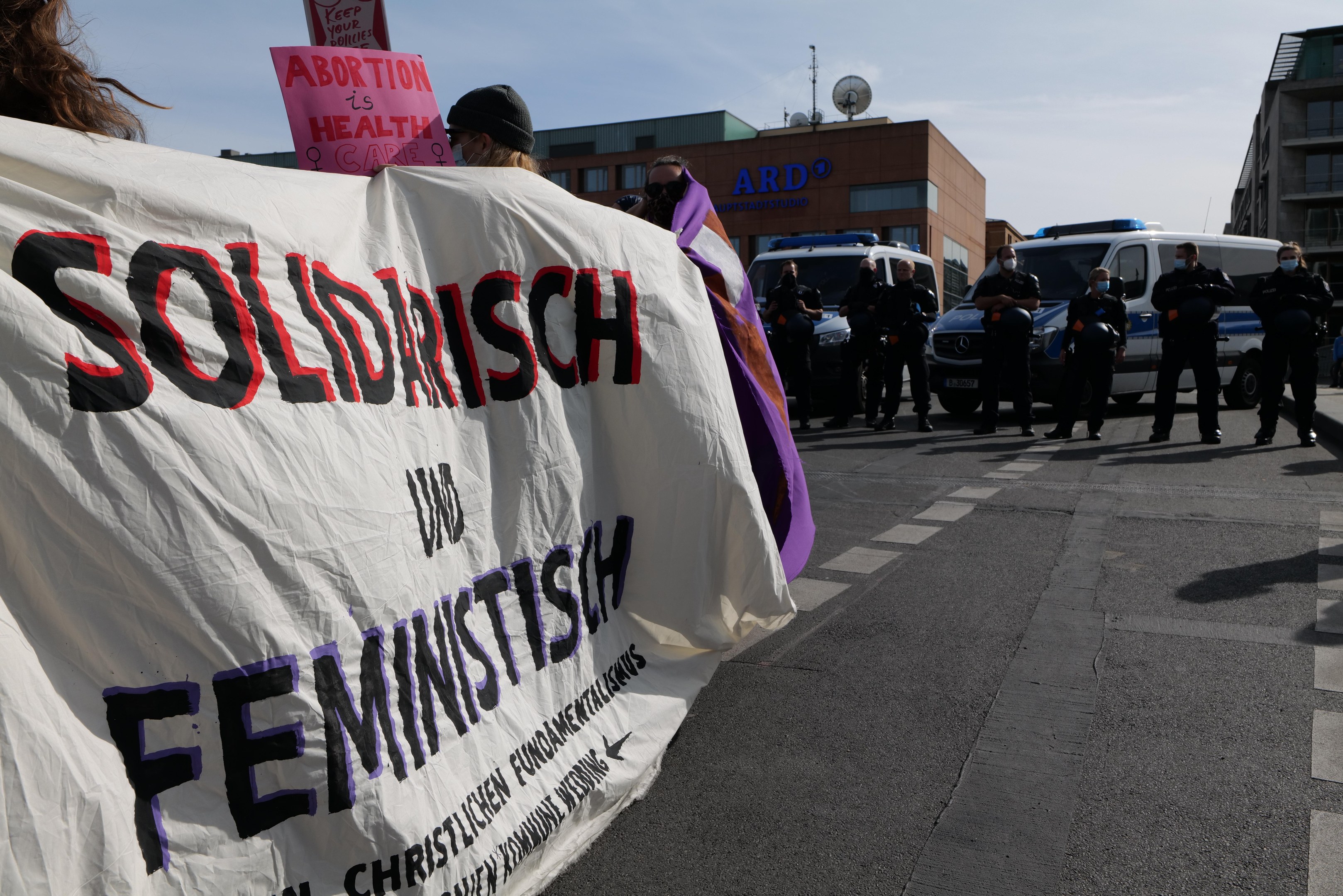 Gruppe von Menschen marschiert die Straße entlang und hält eine 'Solidarität und Feminismus'-Fahne, mit parkenden Fahrzeugen und Gebäuden im Hintergrund unter einem klaren blauen Himmel.