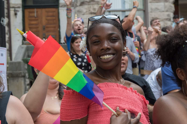 Gruppe von Menschen vor einem Gebäude mit einer Frau in der Mitte, die eine Regenbogenflagge hält, wahrscheinlich bei einer Gay-Pride-Parade