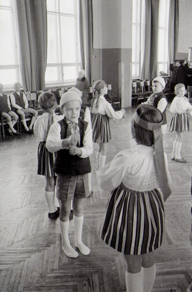 Schwarzes und weißes Foto von Kindern in Schuluniformen, die in einem Raum mit sitzenden Beobachtern im Hintergrund tanzen, Vorhänge an den Fenstern und eine sichtbare Wand.