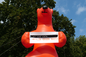 Ein aufblasbarer Bär hält ein Schild mit der Aufschrift "Berlin gegen Nazis" vor Bäumen und einem bewölkten Himmel.