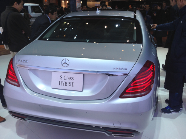 Silberner Mercedes S-Klasse Hybrid auf einer Automesse gezeigt, umgeben von Menschen in verschiedenenfarbigen Kleidern, mit anderen Fahrzeugen und Beleuchtung im Hintergrund.